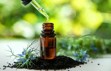 An image of a dark colored vial of oil, and a dropper pouring oil into vial. Vial is placed over black cumin seeds. Herbstonic Hair Oil for Black. 